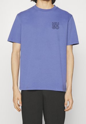 Mężczyzna noszący gładki fioletowy t-shirt z krótkim rękawem z logo tekstowym "HUGO" na górnej lewej części klatki piersiowej, w połączeniu z czarnymi spodniami.