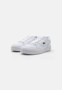 White leather sneakers with perforated toe, flat laces, padded collar, and a small green logo accent on the side. Textured rubber sole.