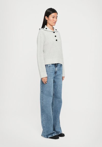 Pull en laine gris avec un large col, des accents floraux noirs et des boutons. Associé à un jean bleu clair à jambes larges et des chaussures noires.