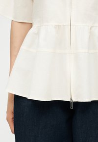 Κοντινό πλάνο ενός ατόμου που φοράει κρεμ peplum τοπ με φερμουάρ μπροστά και σκούρο μπλε παντελόνι, το χέρι ακουμπάει στο πλάι.