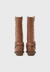 Botas de cuero marrón hasta la rodilla con un acabado ligeramente desgastado, que presentan una correa lateral con herrajes de plata y un tacón bajo apilado.