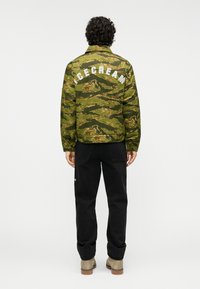 Καμουφλάζ μπουφάν σε πράσινο και καφέ χρώμα με τη λέξη «ICECREAM» σε λευκά γράμματα στην πλάτη. Συνδυασμένο με μαύρο παντελόνι και ανοιχτό καφέ μπότες.