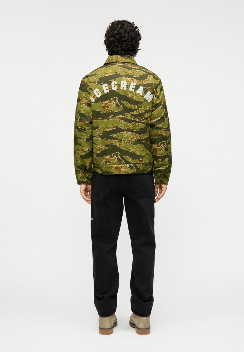 Καμουφλάζ μπουφάν σε πράσινο και καφέ χρώμα με τη λέξη «ICECREAM» σε λευκά γράμματα στην πλάτη. Συνδυασμένο με μαύρο παντελόνι και ανοιχτό καφέ μπότες.