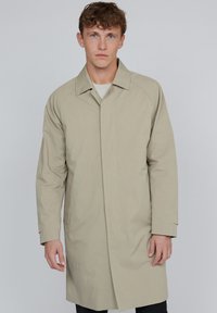 Ung man med ljust brunt hår, iklädd en beige lång kappa över en ljus skjorta, stående mot en enkel grå bakgrund.