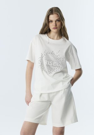 Joven mujer con pantalones cortos blancos y camiseta blanca con el texto "RISE" y un patrón circular de puntos, de pie con una mano en el bolsillo.