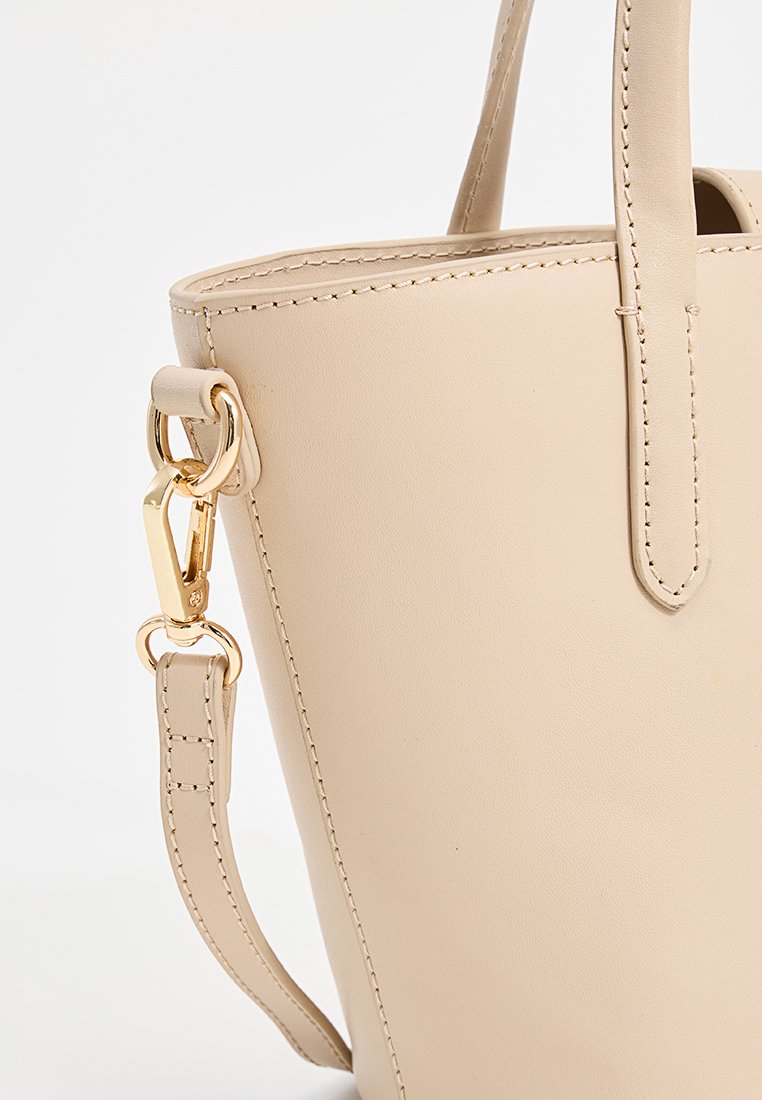 Beige Kunstleder-Shopper mit glatter Textur, ausgestattet mit einem Riemen und goldenen Hardware-Akzenten. Strukturierte Form und minimale Nähdetails.