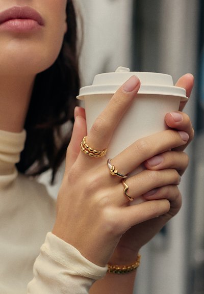 Γυναίκα που κρατάει με τα δύο χέρια ένα λευκό одноχρησιμοποιούμενο ποτήρι καφέ, φορώντας τρία χρυσά δαχτυλίδια και ένα κρεμ μπλουζάκι με μακριά μανίκια.