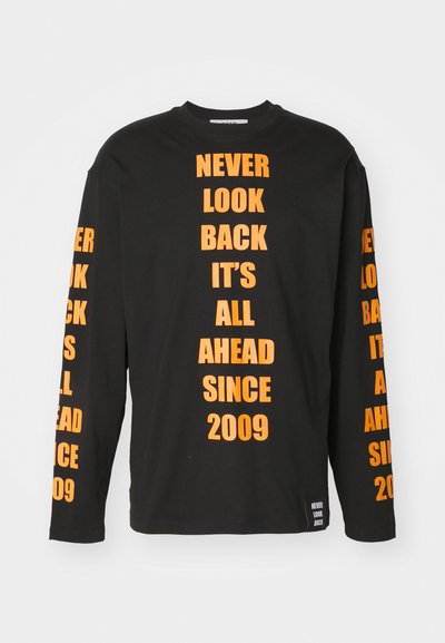 T-shirt noir à manches longues avec du texte orange en gras sur les manches et sur le devant. Le matériau est en coton. Le texte comprend les phrases "NE REGARDEZ JAMAIS EN ARRIÈRE" et "DEPUIS 2009."