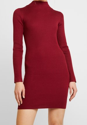 Donna che indossa un vestito aderente a maglia costina a maniche lunghe di colore rosso scuro con scollo alto, che arriva a metà coscia.
