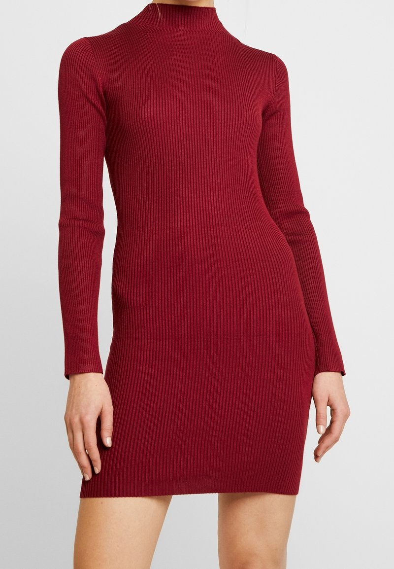 Femme portant une robe près du corps en maille côtelée à manches longues, de couleur rouge foncé, avec un col montant, se terminant à mi-cuisse.