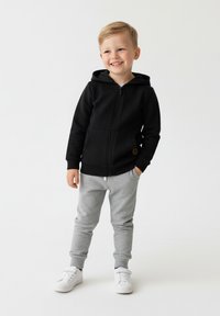 Czarna bluza z kapturem zapinana na zamek, z przednią kieszenią, zestawiona z szarymi spodniami dresowymi. Stylizacja uzupełniona jest białymi trampkami i gładką fakturą.