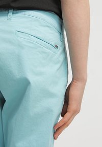 Ljusblå byxor i bomull, med en bakficka som har en subtil metallisk logotypdekoration och en slät textur.