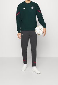 Grön sweatshirt med rosa accenter och Manchester United-logotyp, kombinerad med grå sweatpants som har logotyp och röda detaljer. En vit fotboll hålls.