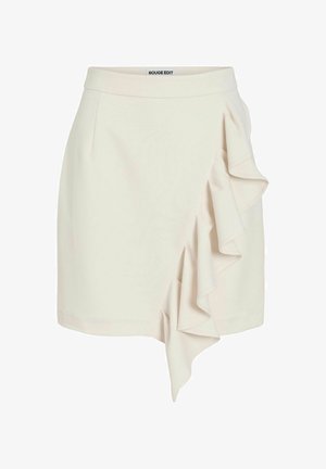 Mini-jupe de couleur crème avec un détail de volant asymétrique sur le côté droit et une ceinture ajustée portant l’étiquette « ROUGE EDIT ».
