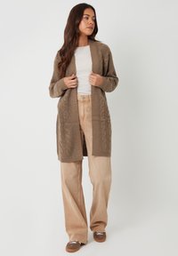 Cárdigan marrón de punto trenzado con un diseño de frente abierto y largo. Combinado con pantalones de pierna ancha beiges claros y zapatillas marrones, sobre un fondo liso.