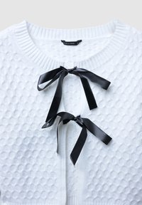 Weißer strukturierter Cardigan mit rundem Ausschnitt, der mit schwarzen Satinschleifen geschlossen wird. Rippbündchen und -saum vervollständigen das Design.