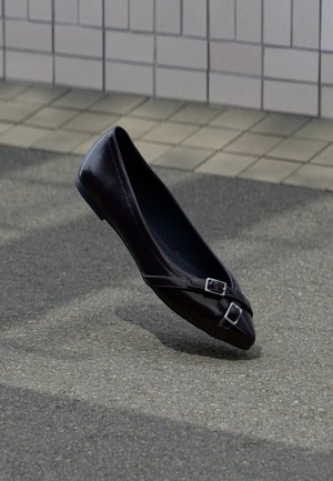 Ballerine plate noire à bout pointu avec deux petites boucles argentées flottant au-dessus de l'asphalte près d'un mur carrelé en arrière-plan.