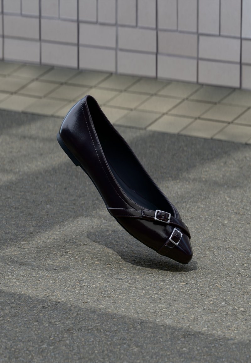 Ballerine plate noire à bout pointu avec deux petites boucles argentées flottant au-dessus de l'asphalte près d'un mur carrelé en arrière-plan.