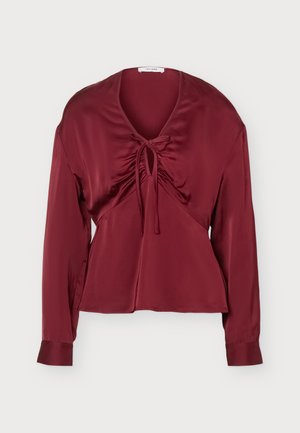 Femme portant un haut bordeaux à manches longues avec un nœud devant et un pantalon noir à jambes larges, debout sur un fond gris clair.