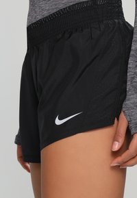 Gros plan sur une personne portant un short de sport noir Nike avec un logo blanc et un haut à manches longues gris, la main reposant sur la cuisse.