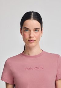 Kortärmad rosa bomullströja med en präglad "Polo Club"-logotyp på framsidan och rundad halsdesign. Slät textur.