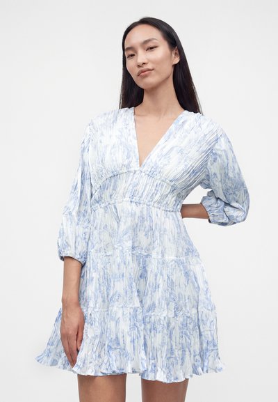 Femme portant une robe à motif floral blanc et bleu clair, col en V, manches longues bouffantes et jupe plissée, posant avec une main sur la hanche.