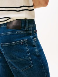 Jeans de ganga azul escuro com uma silhueta ajustada, apresentando um retângulo de couro rotulado "TOMMY HILFIGER" e um pequeno logotipo no bolso.