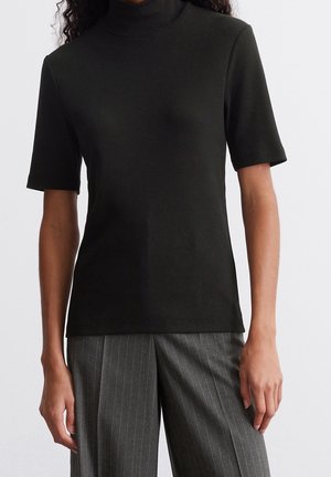 Schwarzes, kurzärmliges Mock-Turtleneck-Oberteil kombiniert mit grauer, breitbeiniger Hose mit Nadelstreifen an einer stehenden Person.