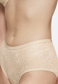Beżowe koronkowe majtki z kwiatowym wzorem, miękką teksturą i gładkim paskiem w talii. Widok z boku podkreśla delikatny design i pokrycie.
