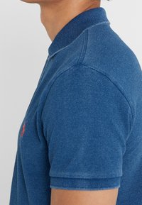 Man som bär en blå texturerad polotröja med en röd broderad logotyp på bröstet, visad från nacken till mitten av överarmen.