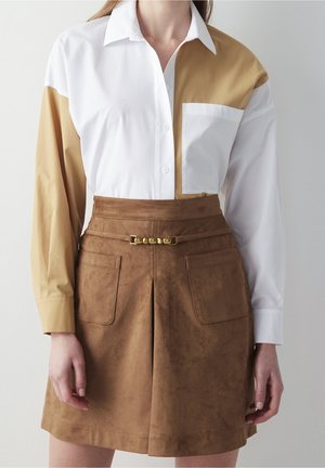 Kvinde iført en hvid og beige skjorte i blokfarver, knappet op og stoppet ned i et brunt ruskindskørt med forlommer og et tyndt bælte.