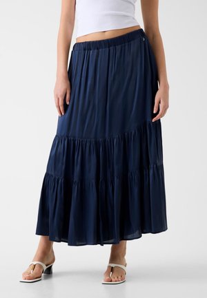 Frau trägt einen marineblauen, mehrstufigen Maxirock mit elastischem Bund, ein weißes kurzes Oberteil und weiße Sandalen mit Absatz.
