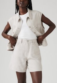 Svetlo bež džins brezrokavnik s sprednjimi gumbi in brezrokavnim dizajnom, v kombinaciji z visokim pasom svetlo bež kratkimi hlačami. Gladka tekstura, sproščen kroj.