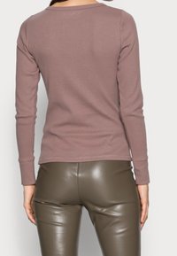Ansicht von hinten einer Person, die ein langärmliges mausfarbiges Oberteil und olivgrüne Lederhosen trägt, und vor einem schlichten hellen Hintergrund steht.