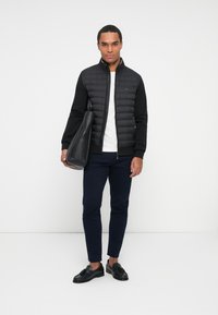 Svart puffjacka med dragkedja fram, svarta ärmar, vit t-shirt, marinblå byxor och svarta loafers. Bär en svart tote-väska.