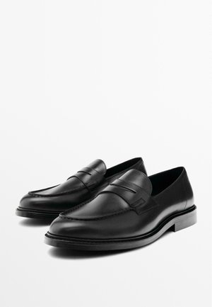 Ett par svarta läder penny loafers med rundade tår och låga staplade klackar på en helt vit bakgrund.