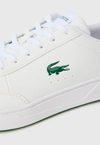 Witte leren sneaker met groen krokodillenlogo, platte veters en geperforeerde details nabij de zool. Textured rubberen buitenzool, modern ontwerp.