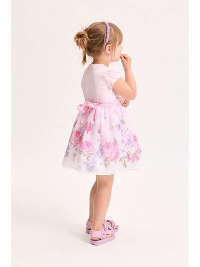 Νεαρή κοπέλα με ροζ φόρεμα με λουλούδια, φιόγκο και ταιριαστά σανδάλια, στέκεται πλευρικά με το χέρι κοντά στο στόμα σε λευκό φόντο.