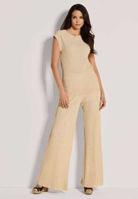 Vrouw die tegen een lichte muur staat, gekleed in een beige, textuurloze mouwloze top en een breedpijpenbroek met zwarte platform sandalen.