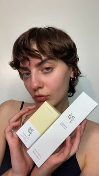 Två förpackningar med hudvårdsprodukter: en gul med en guldfärgad topp och en vit, båda med svart text och varumärke. Hålls upp mot en neutral bakgrund.