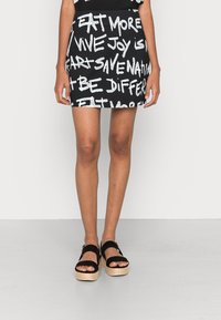 Mini falda negra con texto blanco, con frases en un estilo manuscrito. Combinada con sandalias de plataforma negras y piernas desnudas visibles.