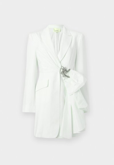 Blazer branco feito à medida com frente drapeada, um grande laço e um broche decorativo prateado. Apresenta mangas compridas e gola com recorte.