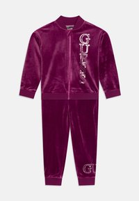 Tuta in velluto di un profondo viola con giacca con zip e pantaloni abbinati. Presenta il logo "GUESS" in strass scintillanti su entrambi i pezzi.