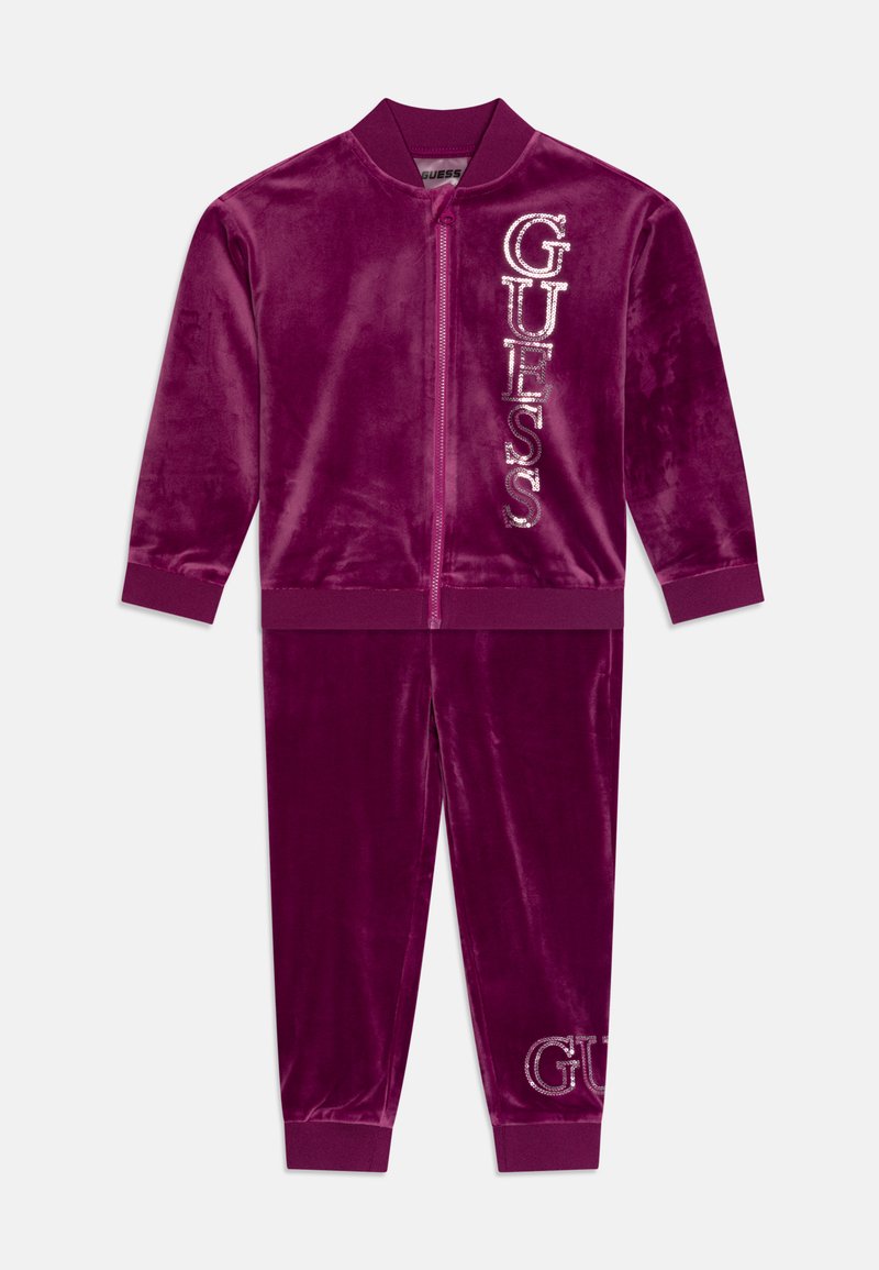 Tuta in velluto di un profondo viola con giacca con zip e pantaloni abbinati. Presenta il logo "GUESS" in strass scintillanti su entrambi i pezzi.