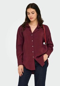 Donna con lunghi capelli scuri che indossa una camicia button-up borgogna larga, jeans scuri e una borsa rossa a spalla, con una mano in tasca.