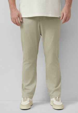 Homme portant un pantalon beige côtelé et des baskets crème, debout devant un fond clair uni, les mains détendues sur les côtés.