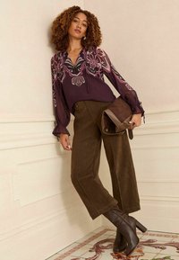 Langärmlige burgunderfarbene Bluse mit floralen Motiven, kombiniert mit braunen Wildleder-Hosen mit weitem Bein und braunen Absatzstiefeln, dazu eine braune Tasche.