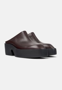 Musta ruskea nahkainen slip-on -kenkä, jossa on paksut mustat pohjat ja leikattu kaarituen tuki, näkyy etukulman näkökulmasta valkoista taustaa vasten.