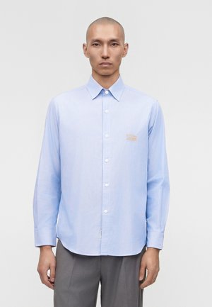 Man die een lichtblauw, langermig overhemd met een klein logo op de borst en een grijze broek draagt, staand tegen een effen achtergrond.