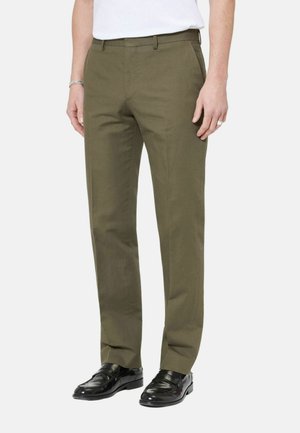 Hombre con pantalones a medida color verde oliva, camisa blanca, mocasines de cuero negro, de pie con las manos relajadas a los lados.